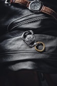 A men's wristwatch with a dark face and brown leather strap rests on a textured black leather surface. Adjacent to the watch is a silver chain adorned with a detailed, lion-shaped accessory attached to a golden ring. The image has a moody, luxurious aesthetic.