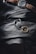 A men's wristwatch with a dark face and brown leather strap rests on a textured black leather surface. Adjacent to the watch is a silver chain adorned with a detailed, lion-shaped accessory attached to a golden ring. The image has a moody, luxurious aesthetic.