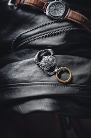A men's wristwatch with a dark face and brown leather strap rests on a textured black leather surface. Adjacent to the watch is a silver chain adorned with a detailed, lion-shaped accessory attached to a golden ring. The image has a moody, luxurious aesthetic.