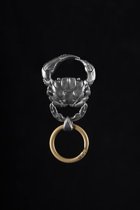 A metallic object resembling a scorpion with spread claws holds a circular, golden ring. The design is intricate, highlighting the polished metal of the scorpion and the contrasting warm tone of the ring.