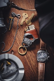 A wooden surface displays a collection of items, including a brown leather-strapped watch, a metallic lighter, two ornate keys with intricate designs, a metal ashtray containing bullet casings, and a decorative keychain with a lion head motif. The setup suggests a theme of vintage or collectible items.