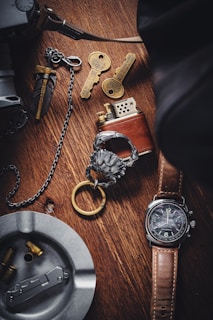 A wooden surface displays a collection of items, including a brown leather-strapped watch, a metallic lighter, two ornate keys with intricate designs, a metal ashtray containing bullet casings, and a decorative keychain with a lion head motif. The setup suggests a theme of vintage or collectible items.