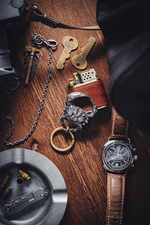 A wooden surface displays a collection of items, including a brown leather-strapped watch, a metallic lighter, two ornate keys with intricate designs, a metal ashtray containing bullet casings, and a decorative keychain with a lion head motif. The setup suggests a theme of vintage or collectible items.