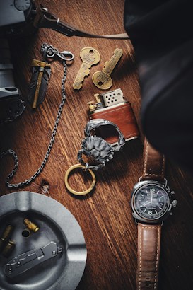 A wooden surface displays a collection of items, including a brown leather-strapped watch, a metallic lighter, two ornate keys with intricate designs, a metal ashtray containing bullet casings, and a decorative keychain with a lion head motif. The setup suggests a theme of vintage or collectible items.