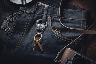 A close-up view of a pair of dark blue denim jeans with distinctive yellow stitching. A metallic key holder attached to the jeans holds two bronze keys and is adorned with a small piece featuring intricate designs. To the left of the jeans, a chain with large, decorative links and several charms, including a feather, are visible. At the bottom right corner, a brown camera bag with a strap is partially visible.