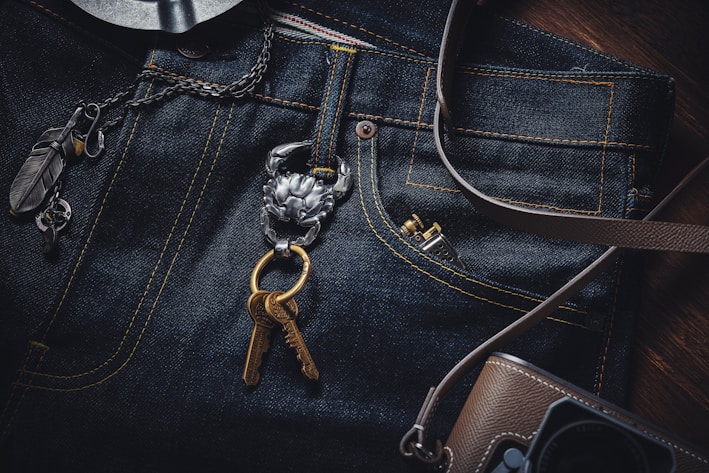 A close-up view of a pair of dark blue denim jeans with distinctive yellow stitching. A metallic key holder attached to the jeans holds two bronze keys and is adorned with a small piece featuring intricate designs. To the left of the jeans, a chain with large, decorative links and several charms, including a feather, are visible. At the bottom right corner, a brown camera bag with a strap is partially visible.