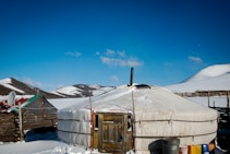 A circular, tent-like structure known as a yurt is situated in a snowy landscape. The yurt is made from a white material and has a small chimney protruding from its roof. Snow-covered hills and a clear blue sky fill the background. A wooden fence and some colorful containers are visible nearby, and a satellite dish is affixed to an adjacent structure.