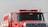 A bright red fire truck with its front facing the viewer, featuring a prominent grille, side mirrors, and emergency lights on top. The truck appears robust and ready for action, displaying various control panels and equipment storage areas on its side.