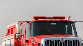 A bright red fire truck with its front facing the viewer, featuring a prominent grille, side mirrors, and emergency lights on top. The truck appears robust and ready for action, displaying various control panels and equipment storage areas on its side.