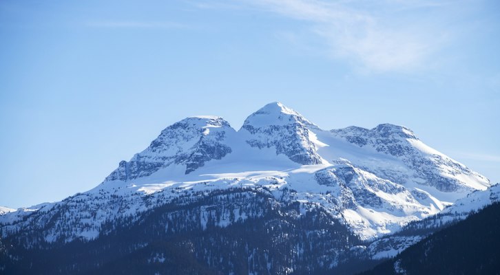 Mount Begbie's twin snowy summits and glacier rising above dark forest under a deep blue sky, Revelstoke