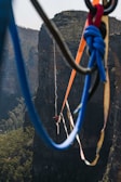 A person is walking on a highline stretched between two cliffs. The scene captures the breathtaking height and the rugged natural landscape of a canyon with lush greenery below. Vibrant climbing ropes of various colors are visible in the foreground, adding a dynamic element to the composition.