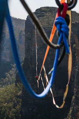 A person is walking on a highline stretched between two cliffs. The scene captures the breathtaking height and the rugged natural landscape of a canyon with lush greenery below. Vibrant climbing ropes of various colors are visible in the foreground, adding a dynamic element to the composition.