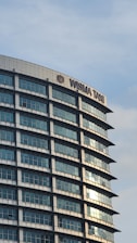 A modern, curved building facade with reflective glass windows and a sign that reads 'WISMA TANI' near the top. The building features a grid pattern due to its window design, and the sky is partly cloudy with a light blue hue.