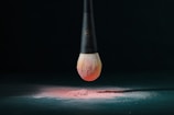 A makeup brush with a black handle is suspended above a surface, covered with a fine layer of pink powder. The bristles of the brush are dipped in the same pink powder, creating a gradient effect. The setting is dark, emphasizing the brush and powder.