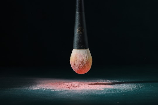 A makeup brush with a black handle is suspended above a surface, covered with a fine layer of pink powder. The bristles of the brush are dipped in the same pink powder, creating a gradient effect. The setting is dark, emphasizing the brush and powder.