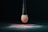 A makeup brush with a black handle is suspended above a surface, covered with a fine layer of pink powder. The bristles of the brush are dipped in the same pink powder, creating a gradient effect. The setting is dark, emphasizing the brush and powder.