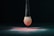 A makeup brush with a black handle is suspended above a surface, covered with a fine layer of pink powder. The bristles of the brush are dipped in the same pink powder, creating a gradient effect. The setting is dark, emphasizing the brush and powder.