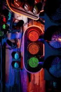 A display of multiple traffic lights mounted on a wall, with a central focus on one set showing a red and green light. The scene is illuminated by colored lighting, casting a mix of blue, purple, and warm orange tones over the area. The background includes an industrial-style wall with visible wires and mounts.