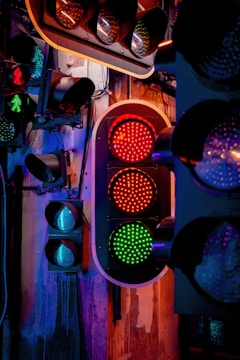 A display of multiple traffic lights mounted on a wall, with a central focus on one set showing a red and green light. The scene is illuminated by colored lighting, casting a mix of blue, purple, and warm orange tones over the area. The background includes an industrial-style wall with visible wires and mounts.