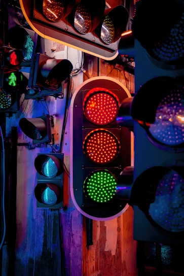 A display of multiple traffic lights mounted on a wall, with a central focus on one set showing a red and green light. The scene is illuminated by colored lighting, casting a mix of blue, purple, and warm orange tones over the area. The background includes an industrial-style wall with visible wires and mounts.