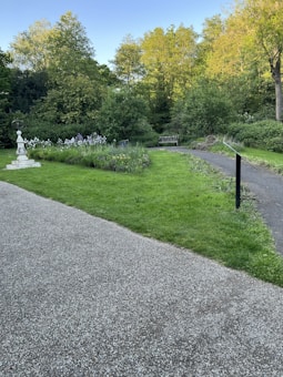 A serene garden scene featuring a pathway and manicured lawns surrounded by lush green trees and bushes. In the foreground, there is a gravel path next to a grassy area with various plants and flowers. A small white stone structure adds a decorative element, and a wooden bench is nestled within the greenery.
