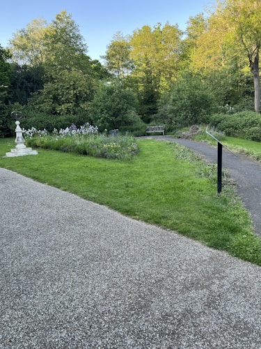 A serene garden scene featuring a pathway and manicured lawns surrounded by lush green trees and bushes. In the foreground, there is a gravel path next to a grassy area with various plants and flowers. A small white stone structure adds a decorative element, and a wooden bench is nestled within the greenery.