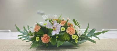 A floral arrangement consisting of various flowers such as white and peach blossoms, set in a green oblong vase. The foreground displays lush green leaves and ferns enhancing the intricate textures. The arrangement sits on a beige patterned surface with a blurred neutral background, giving it a serene and elegant appearance.