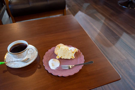Close-up of a steaming cup of coffee next to a slice of delicate pink-frosted cake on a rustic wooden table.