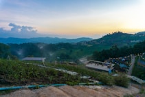 The image depicts a tranquil hilly landscape at sunset, with layers of lush greenery and cultivated fields stretching into the distance. Small structures are scattered throughout the hills, some with roofs visible. A winding pathway bordered by lights leads into the scene, creating a charming, serene atmosphere. Mist rises gently from the irrigated fields, adding a mystical quality to the landscape.