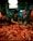 A woman wearing a dark apron is cutting meat at a butcher's stall. She is surrounded by various cuts of raw meat laid out on a red surface. Overhead lighting creates a warm ambiance, and hanging meat is visible in the background.