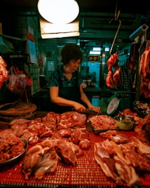 A woman wearing a dark apron is cutting meat at a butcher's stall. She is surrounded by various cuts of raw meat laid out on a red surface. Overhead lighting creates a warm ambiance, and hanging meat is visible in the background.