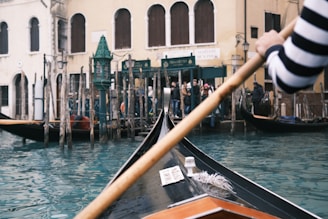 A gondola navigates through a canal, steered by a person wearing a black and white striped shirt. The water is a vibrant turquoise blue, and the background features historic Venetian architecture, including buildings with arched windows. Several wooden poles line the canal's edge, and there is a group of people standing on the shore.
