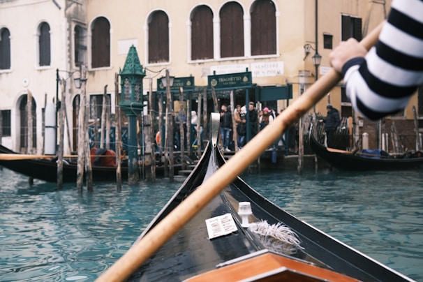 A gondola navigates through a canal, steered by a person wearing a black and white striped shirt. The water is a vibrant turquoise blue, and the background features historic Venetian architecture, including buildings with arched windows. Several wooden poles line the canal's edge, and there is a group of people standing on the shore.