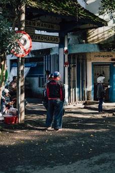 A group of people gather near a street intersection under a leafy canopy. One person stands wearing a helmet, while another sits near a red plastic basket on the pavement. Signage with various place names is visible, situated above the group. Shadows and sunlight are cast across the scene, creating a patchwork of light on the ground.