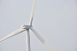 A wind turbine with large white blades set against a pale sky. The turbine has a company logo on the central hub.