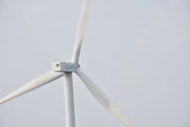 A wind turbine with large white blades set against a pale sky. The turbine has a company logo on the central hub.