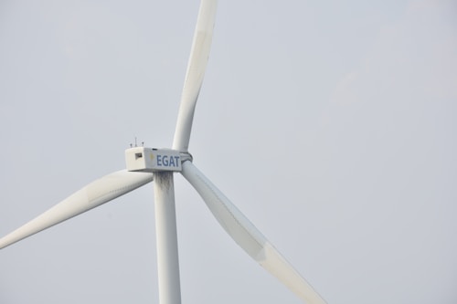 A wind turbine with large white blades set against a pale sky. The turbine has a company logo on the central hub.