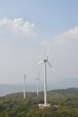 Three large wind turbines are positioned on a lush green hillside, surrounded by dense vegetation. In the background, a hazy view of water and distant mountains is visible under a sky filled with soft, fluffy clouds.