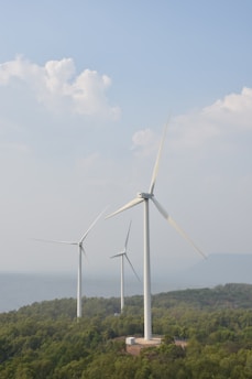 Three large wind turbines are positioned on a lush green hillside, surrounded by dense vegetation. In the background, a hazy view of water and distant mountains is visible under a sky filled with soft, fluffy clouds.