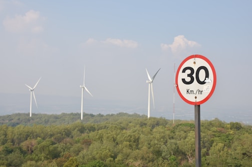 A landscape with several large wind turbines in the background standing tall above a dense green forest. In the foreground, a speed limit sign is visible, indicating a maximum speed of 30 km/h. The sky is clear with a few scattered clouds.