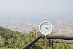 A large thermometer is mounted on a metal railing overlooking a scenic landscape with fields and hills in the distance. The sky is overcast with a slight haze, giving the scene a muted and calm appearance. Vegetation is visible around the perimeter, including bushes and trees.