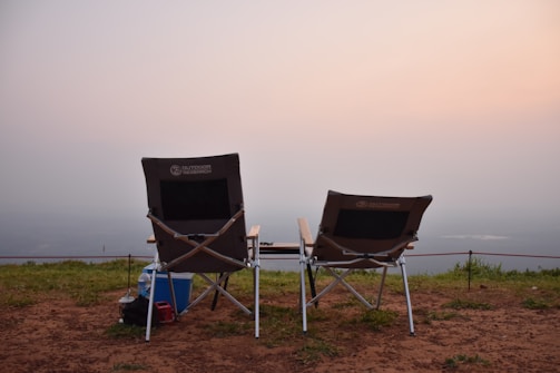 Two foldable camping chairs are placed on a grassy and earthy cliff edge, overlooking a foggy landscape at dawn or dusk. A cooler and some bags are positioned next to the chairs, suggesting preparation for a relaxed outdoor experience. The sky is softly lit with pastel hues, creating a serene and peaceful atmosphere.