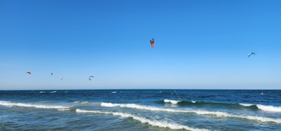 Colorful kitesurfing kites float in a clear blue sky above a serene ocean, with gentle waves rolling towards the sandy shore.