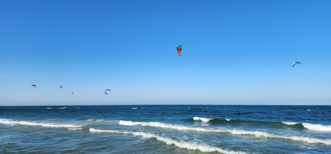 Colorful kitesurfing kites float in a clear blue sky above a serene ocean, with gentle waves rolling towards the sandy shore.