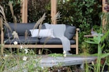 A comfortable outdoor seating arrangement amidst lush greenery features a dark gray cushioned sofa with a light gray blanket and a white pillow. Tall grasses and delicate white flowers occupy the foreground, adding a natural and serene atmosphere. A small table with drinks can be seen on one side, enhancing the cozy garden setting.