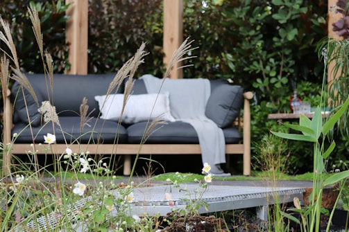 A comfortable outdoor seating arrangement amidst lush greenery features a dark gray cushioned sofa with a light gray blanket and a white pillow. Tall grasses and delicate white flowers occupy the foreground, adding a natural and serene atmosphere. A small table with drinks can be seen on one side, enhancing the cozy garden setting.