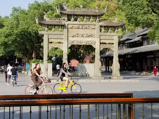A group of people are cycling in a park-like setting with a traditional stone archway in the background. The area is lush with green trees, and there are traditional style buildings to the right. Some people are walking, and the scene conveys a relaxed, leisurely atmosphere.