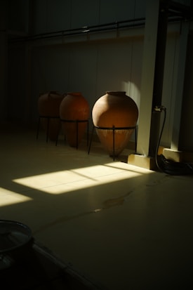Three large terracotta pots are placed on metal stands in a dimly lit indoor space. Sunlight streams through a window, casting a geometric pattern of light and shadow on the floor. The setting appears industrial or utilitarian, with a pipe running along one side of the room.