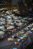 An assortment of colorful stone or gem pendants displayed on a dark cloth background. The pendants are arranged in neat rows and vary in color and pattern, including shades of blue, orange, green, and earth tones. Soft natural light creates a play of shadows, enhancing the visual appeal of the stones.