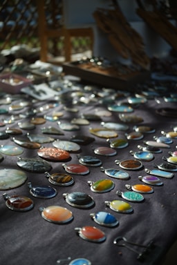 An assortment of colorful stone or gem pendants displayed on a dark cloth background. The pendants are arranged in neat rows and vary in color and pattern, including shades of blue, orange, green, and earth tones. Soft natural light creates a play of shadows, enhancing the visual appeal of the stones.